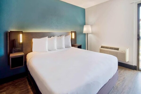 A hotel room with a large bed, two bedside tables, a nightstand, a wall-mounted lamp, and a window with blinds.