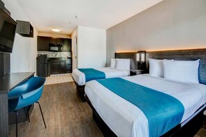 A hotel room with two beds, a desk, a chair, a kitchenette, and a TV.