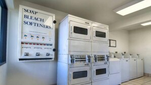 Een wasserette met gestapelde wasmachines en drogers, een zeep-, bleek- en verzachterdispenser, en een bordje waarop de beschikbare soorten wasmiddel staan aangegeven.