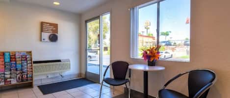 A hotel lobby with a reception desk, a seating area with two chairs and a table, a magazine rack, and a window with a view of the outside.