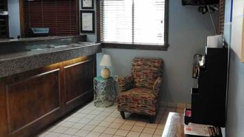 A small kitchen with a tiled floor, a wooden counter, a patterned armchair, a small table with a lamp, and a television mounted on the wall.