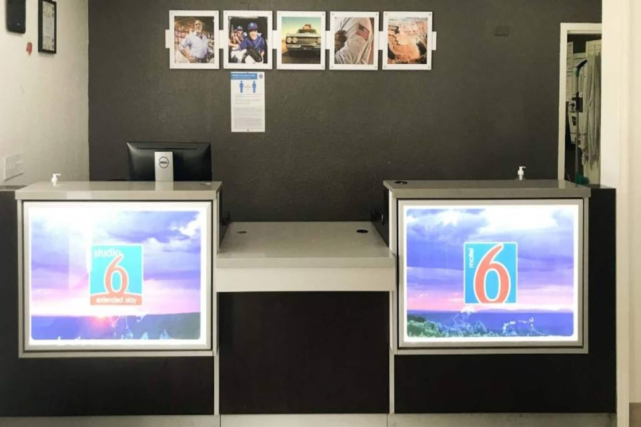 A reception desk with two monitors displaying the number 6, a bulletin board with photos, and a dark-colored wall.