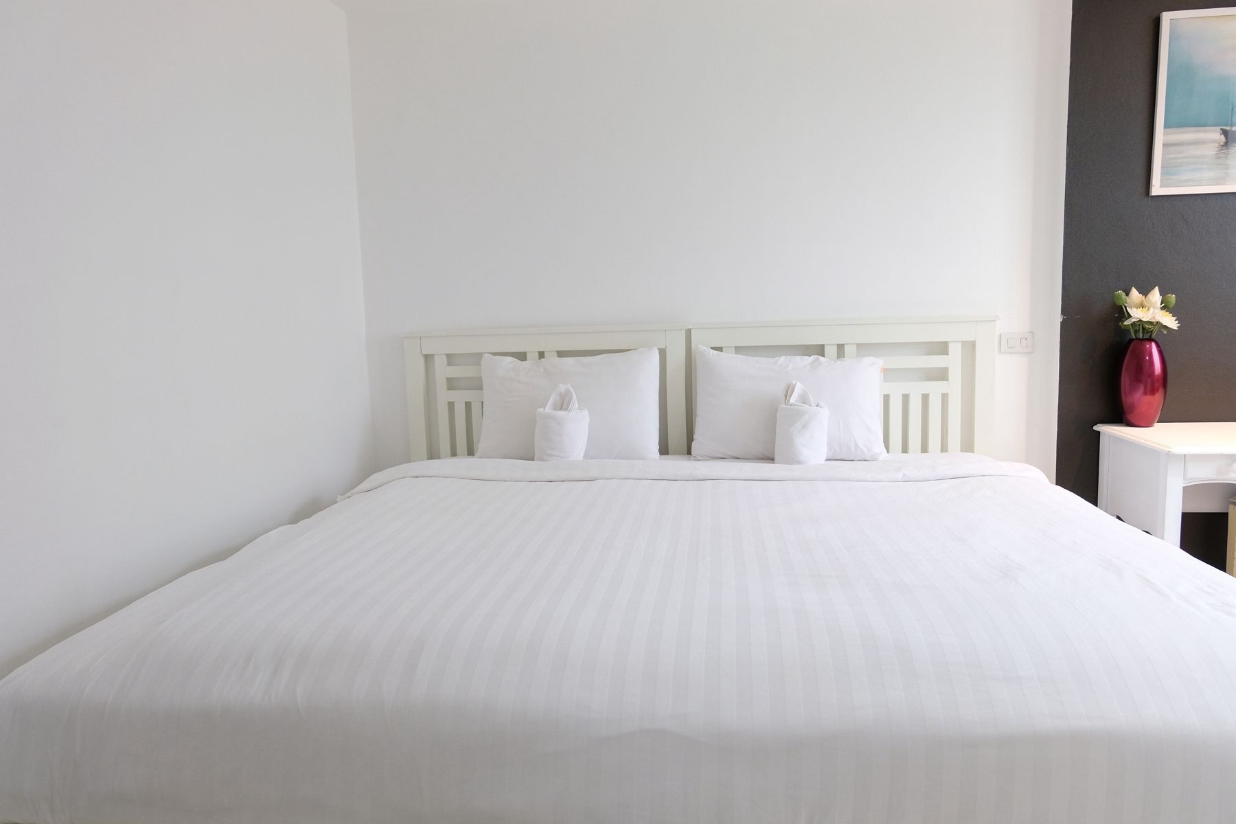Una cama individual con ropa de cama y almohadas blancas, un cabecero blanco y una mesita de noche con un florero.