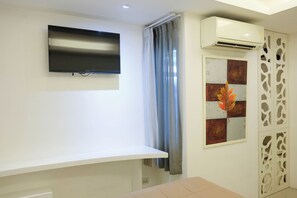 A room with a mounted television, a white wall, a window with curtains, and an air conditioning unit. - Baanpoon Apartment (Koh Samui)