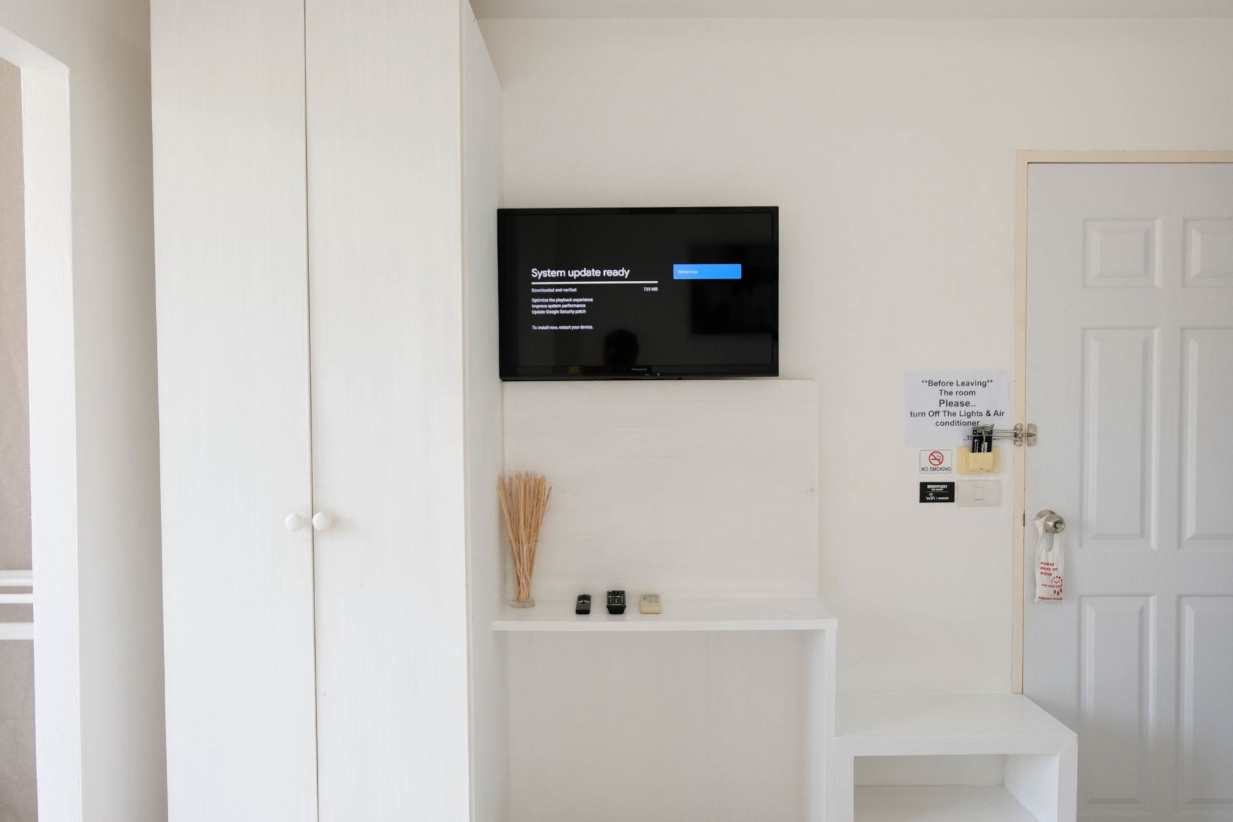 Una habitación con un televisor montado en la pared mostrando un mensaje de actualización del sistema. Hay una puerta blanca con un letrero solicitando apagar las luces y el aire acondicionado antes de salir.