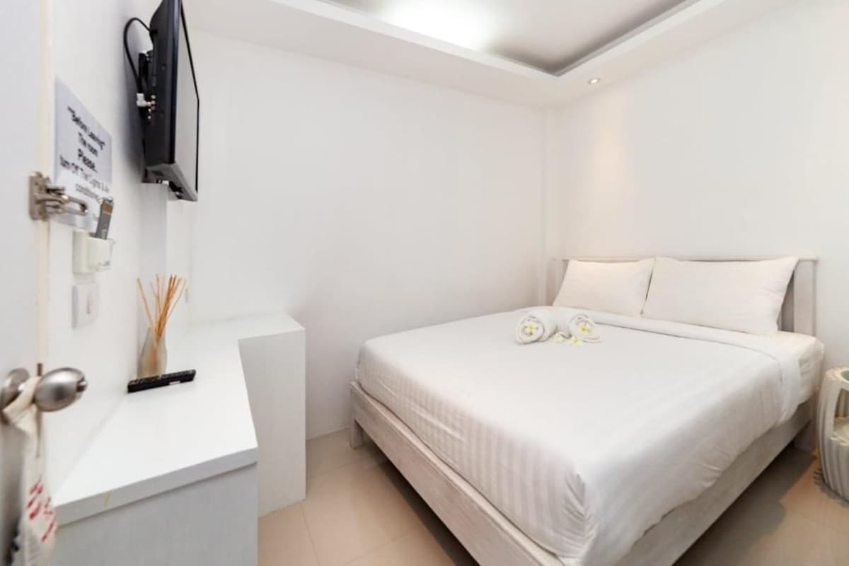 Moderni hotellihuone, jossa on sänky, työpöytä, televisio ja pieni maljakko, jossa on koristeellisia oksia.