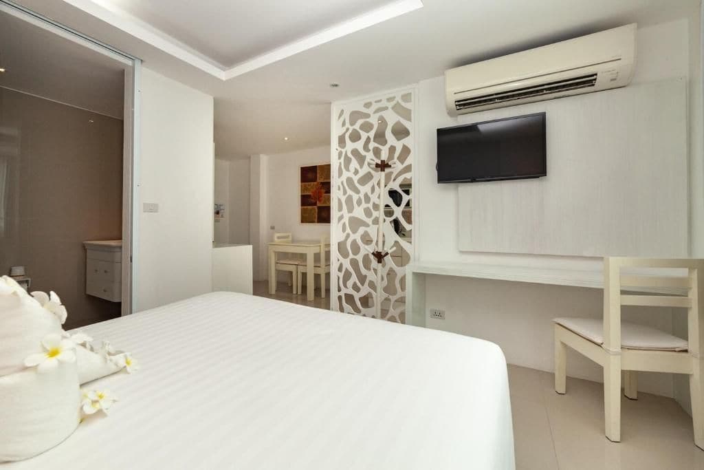 A hotel room with a bed, a TV mounted on the wall, an air conditioning unit, and a small seating area with a chair.
