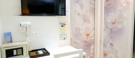 A hotel room with a flat-screen TV mounted above a desk, a hair dryer, a digital safe, and a wall with floral wallpaper.