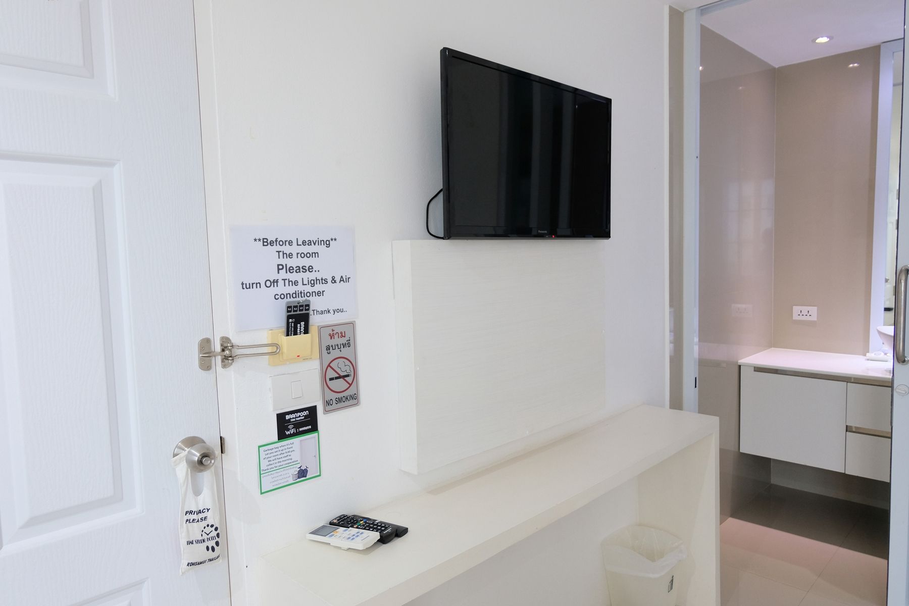 A hotel room with a white door, a mounted television, a small table with a remote control, and various signs including a no-smoking sign and a privacy notice.