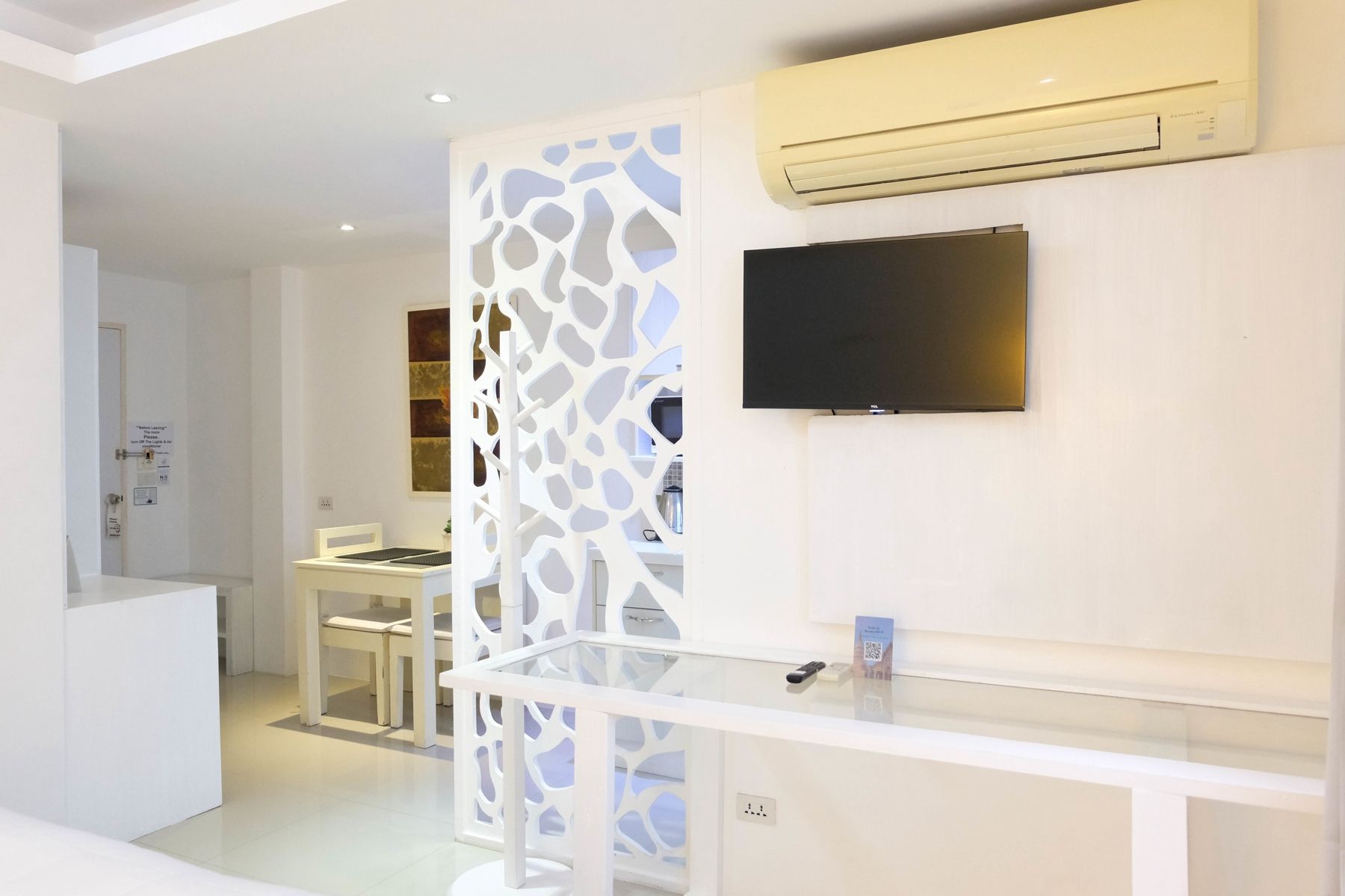 A modern hotel room with a white reception desk, a flat-screen TV mounted on the wall, and an air conditioning unit above it.