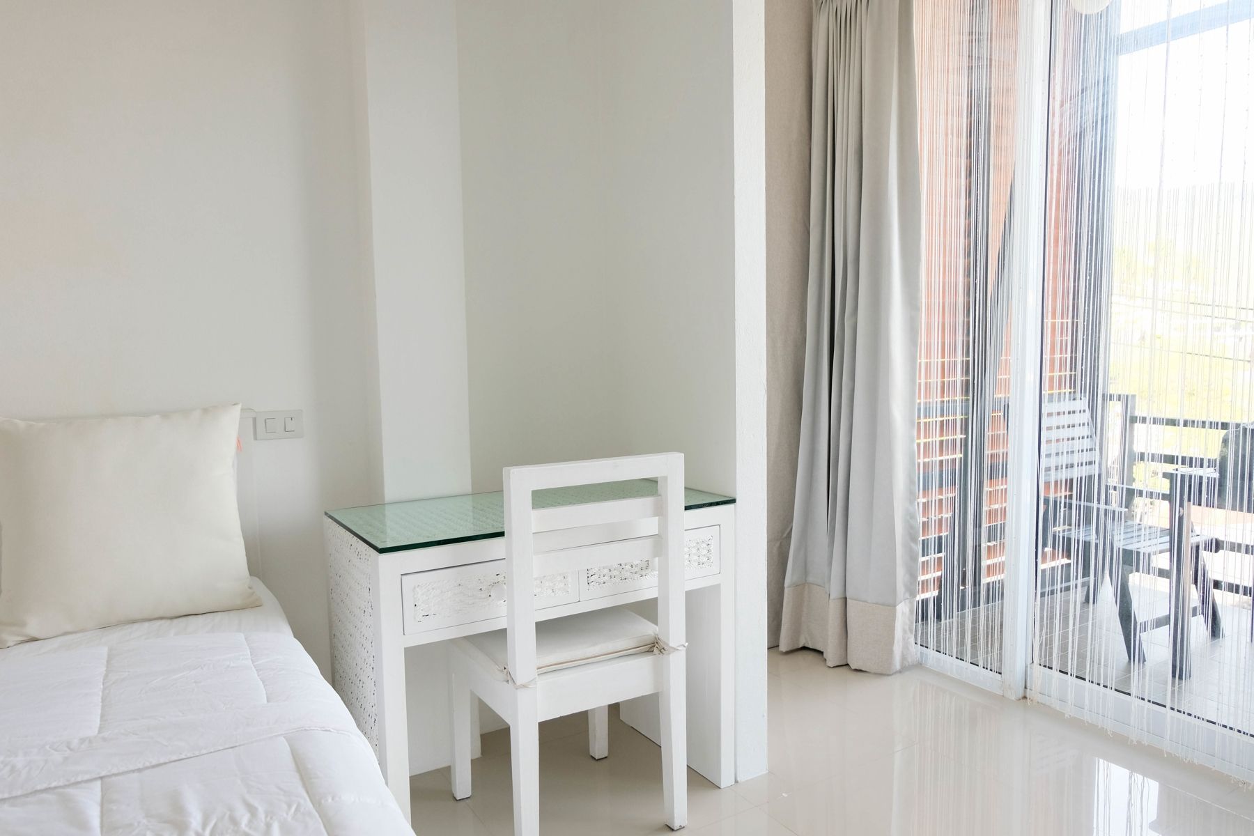 Habitación de hotel con cama, un escritorio blanco con silla y vistas a edificios a través de cortinas translúcidas.