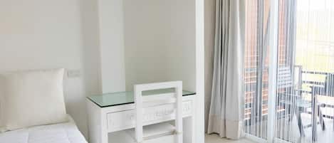 Habitación de hotel con cama, un escritorio blanco con silla y vistas a edificios a través de cortinas translúcidas.