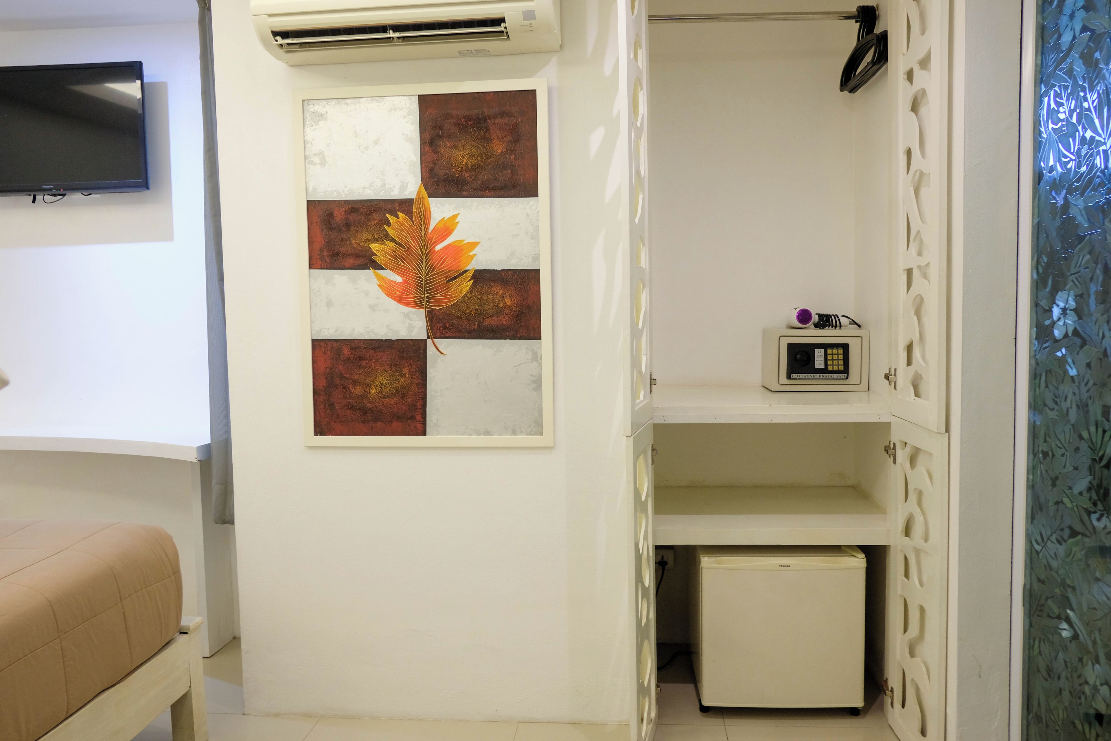 A room with a bed, a wall-mounted TV, an air conditioner, a framed artwork, a small shelf with a device, and a refrigerator.