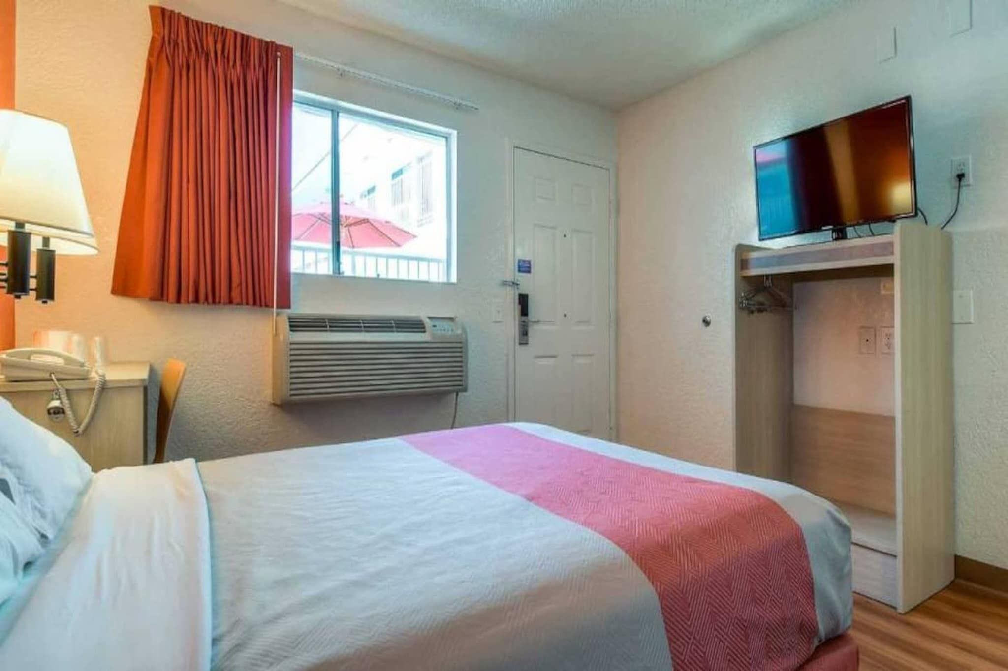 Habitació d'hotel amb un llit, una televisió, un aire condicionat, una taula de nit amb un telèfon i una finestra amb cortines.