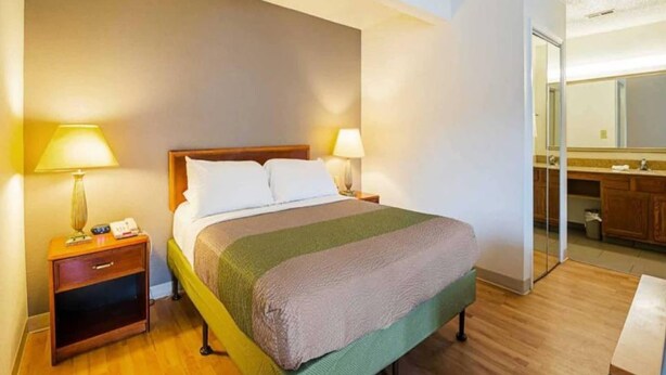 Une chambre d’hôtel avec un lit, des tables de chevet, une lampe, un miroir et une salle de bain visible à travers une porte ouverte.