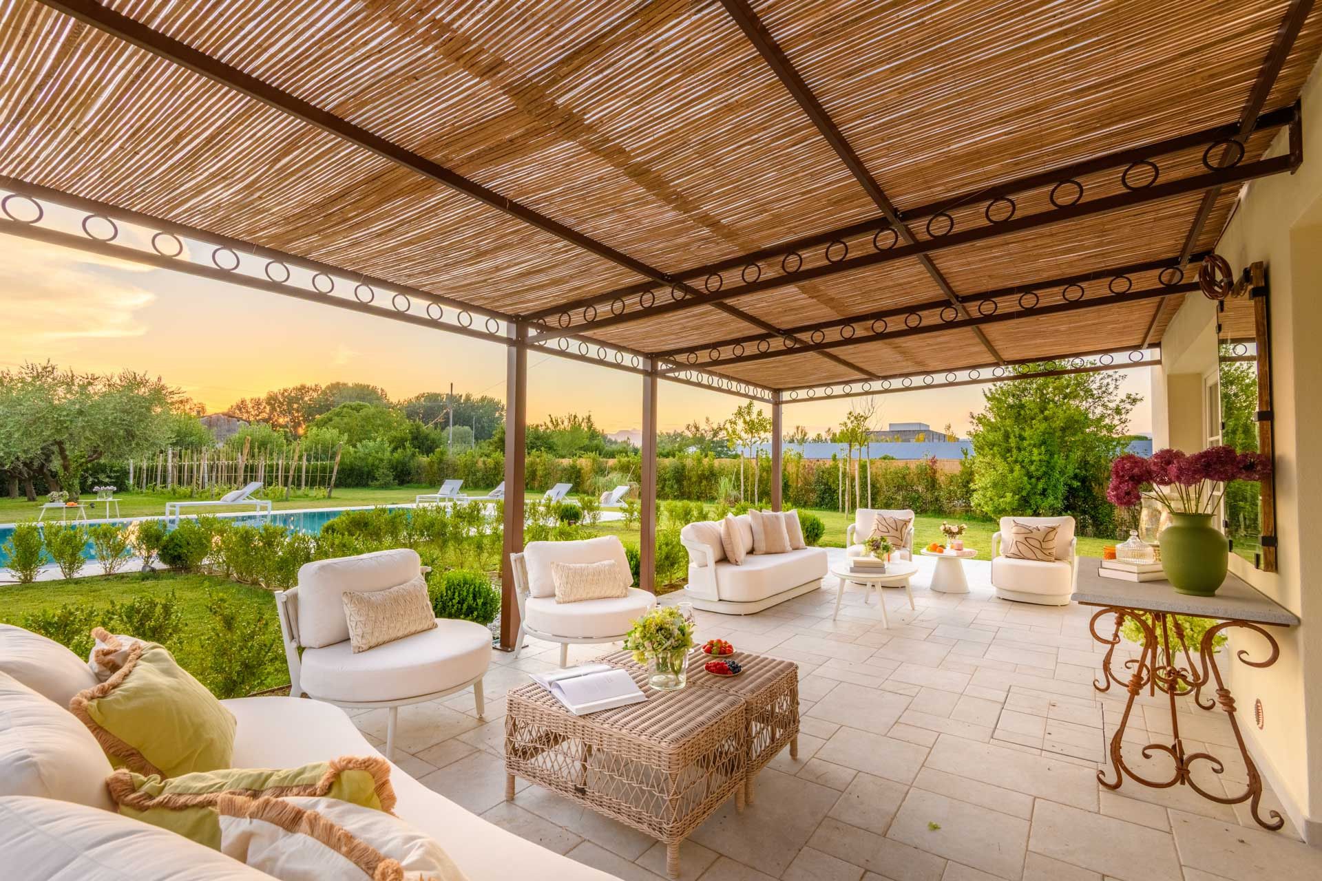 A covered outdoor seating area with white sofas and a wicker coffee table, overlooking a swimming pool and lush greenery.