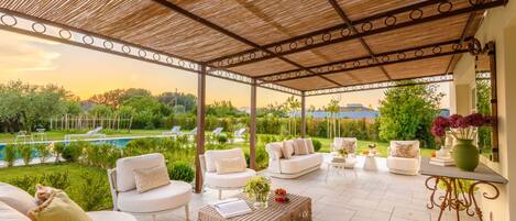 A covered outdoor seating area with white sofas and a wicker coffee table, overlooking a swimming pool and lush greenery.