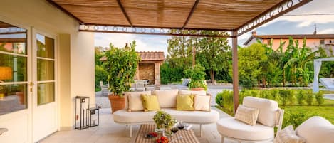 A covered outdoor seating area with wicker furniture, a table with fruit and wine glasses, and potted plants.