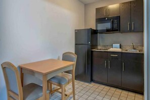 A small kitchen with a refrigerator, microwave, and dark cabinets. There is a wooden table with two chairs.