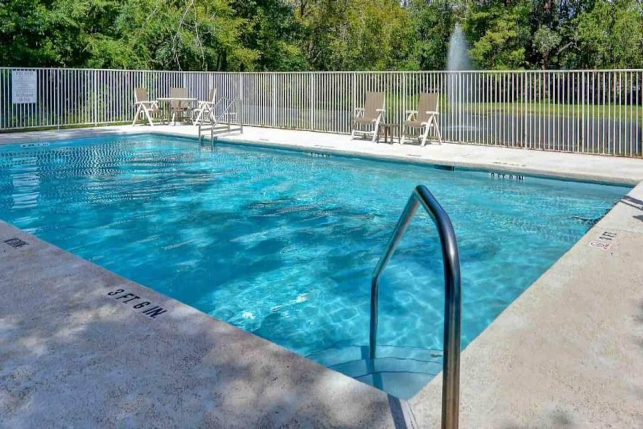Une piscine rectangulaire avec un escalier en métal sur un côté, entourée d’une clôture et bordée d’arbres en arrière-plan.