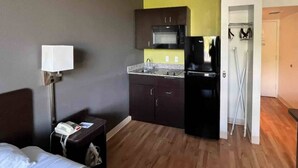 Una habitación de hotel con cama, escritorio con lámpara, kitchenette con microondas y refrigerador, y puerta que da a un baño.