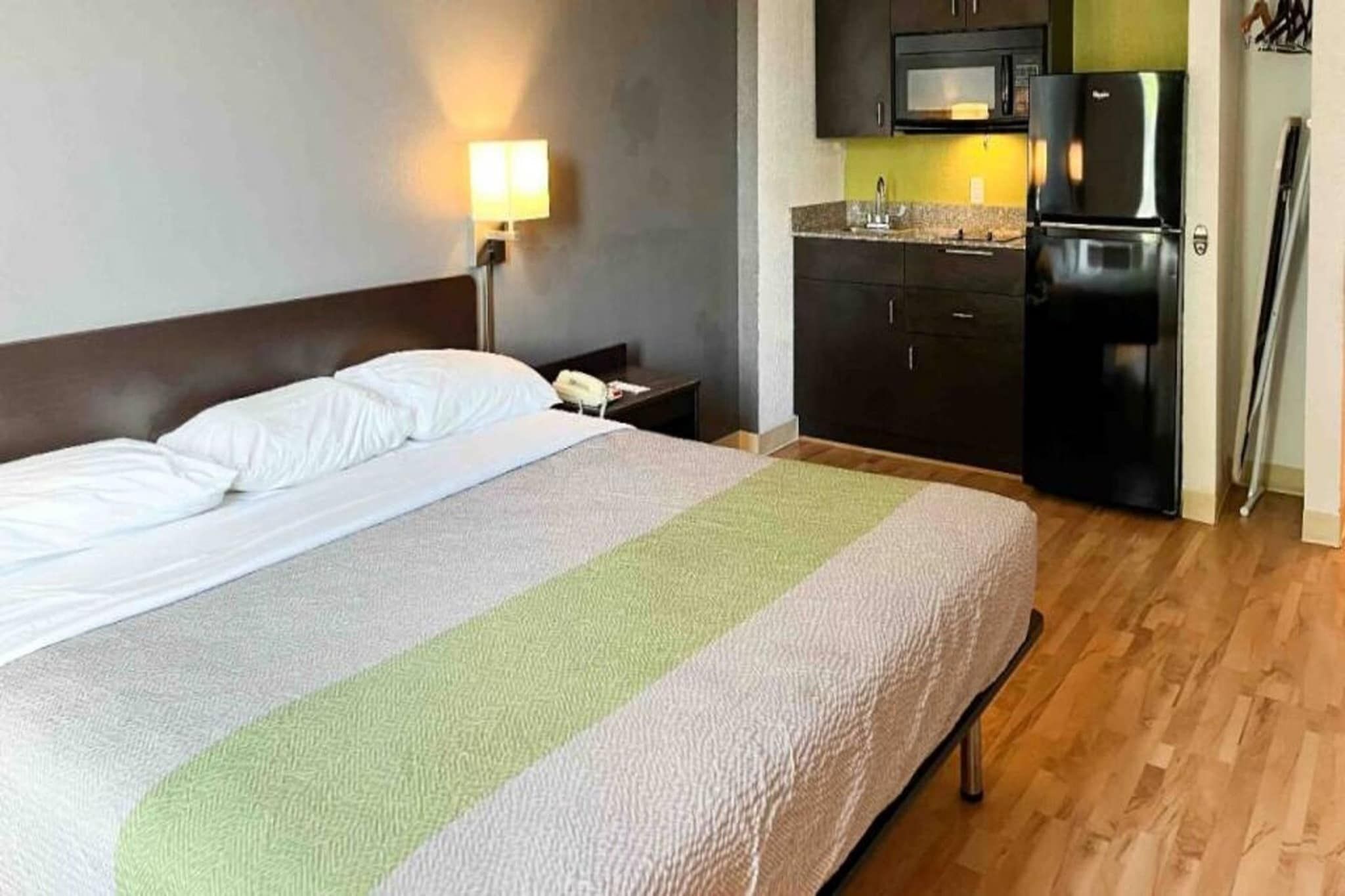 Ett hotellrum med en säng, ett nattduksbord, en lampa, ett litet kök med mikrovågsugn och kylskåp samt ett trägolv.