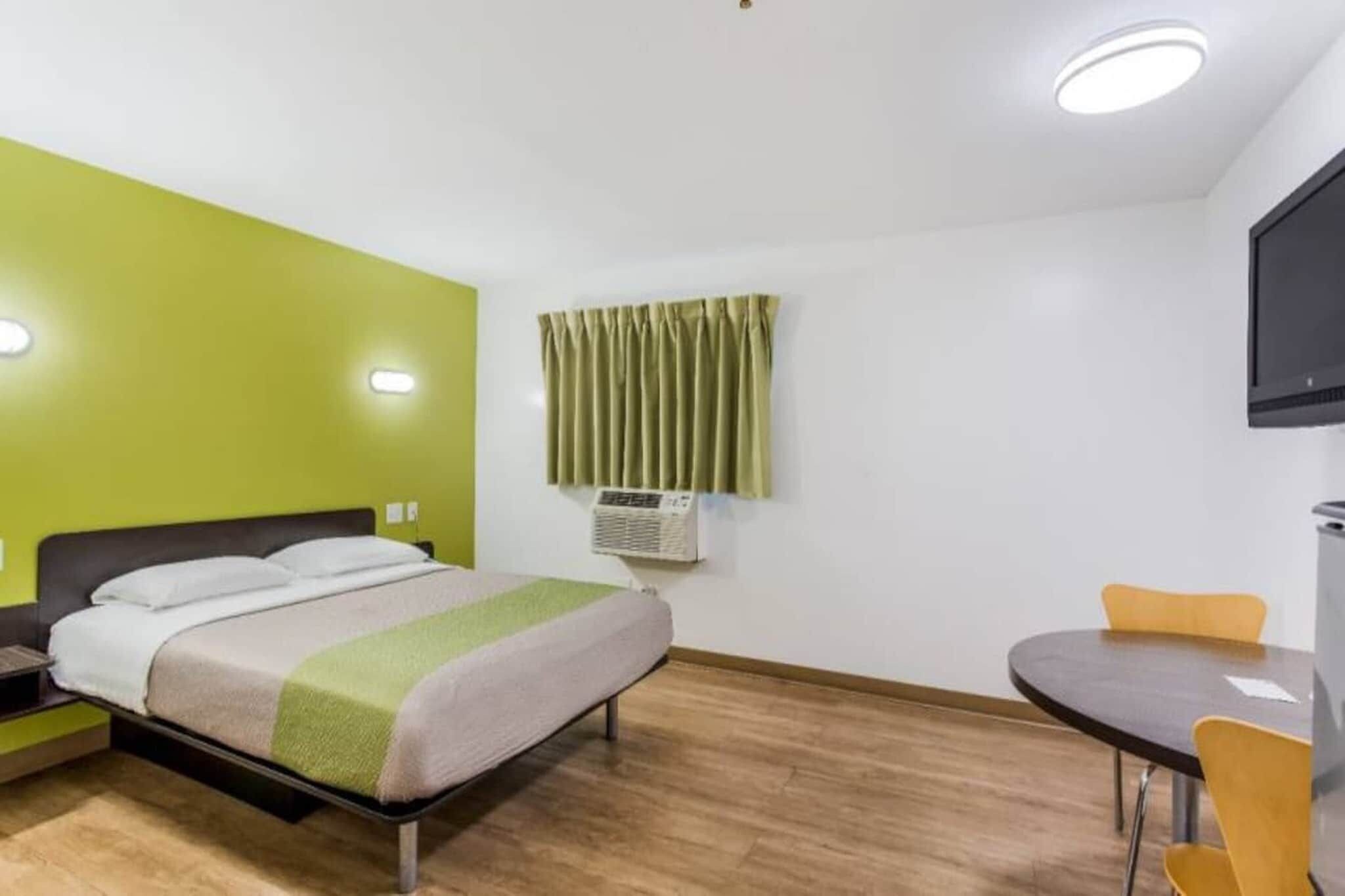 A hotel room with a bed, a small table, a chair, an air conditioning unit, and a television.