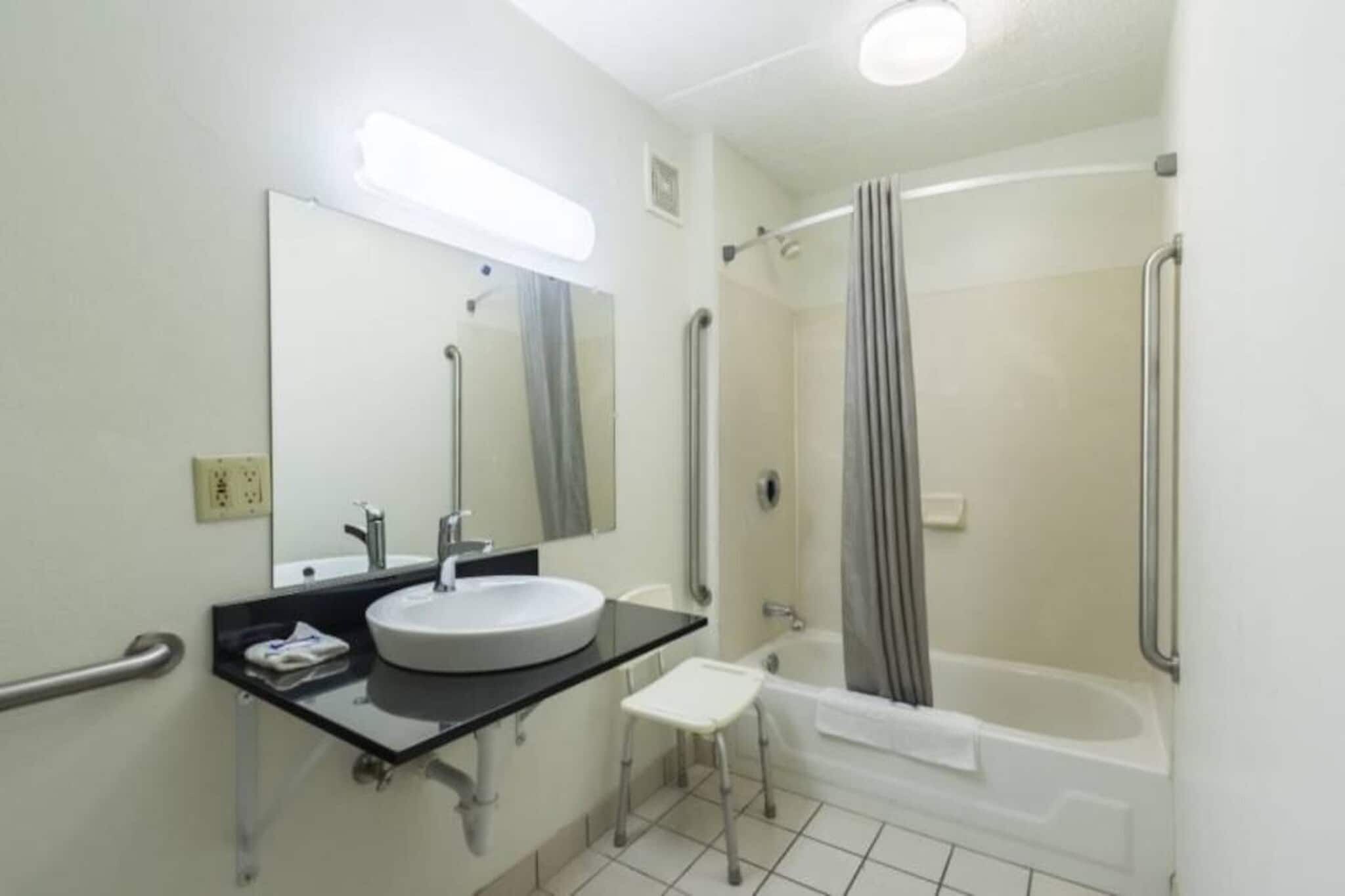 A bathroom with a sink, mirror, handrails, a shower, and a small stool.