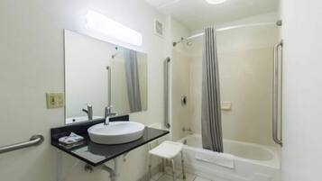 A bathroom with a sink, mirror, handrails, a shower, and a small stool.