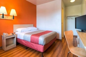 Quarto de hotel com cama, mesa de cabeceira, luminária, cadeira e televisão.