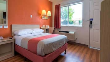 A hotel room with a bed, bedside table, lamp, window, and an air conditioning unit.
