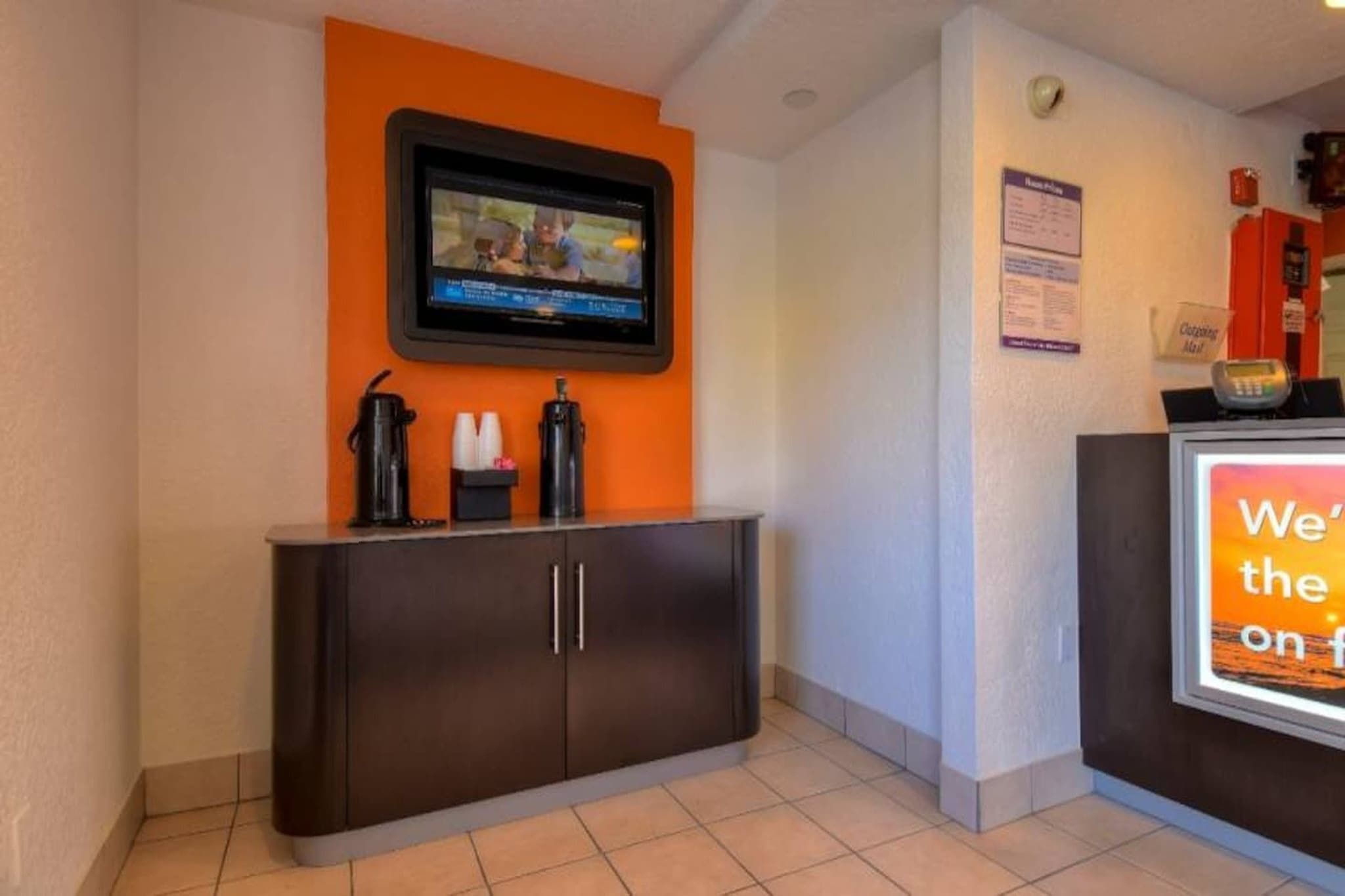 Una zona de recepció amb un televisor de pantalla plana fixat a la paret, un taulell de recepció de color fosc i un senyal amb text parcialment visible.