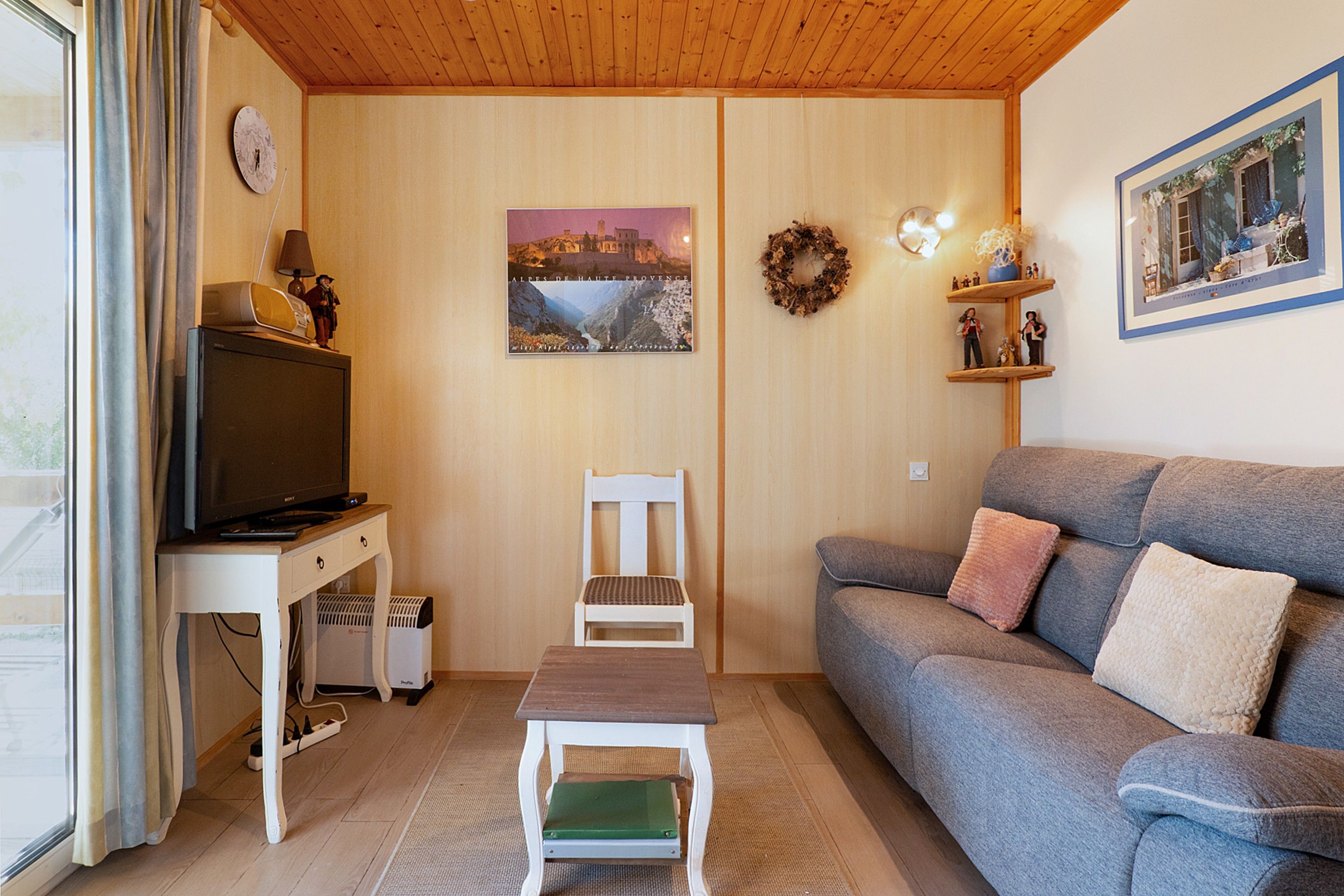 Una sala de estar con un televisor sobre un mueble blanco, un sofá gris con cojines, una pequeña silla blanca, una mesa de madera y una imagen de los Alpes en la pared.