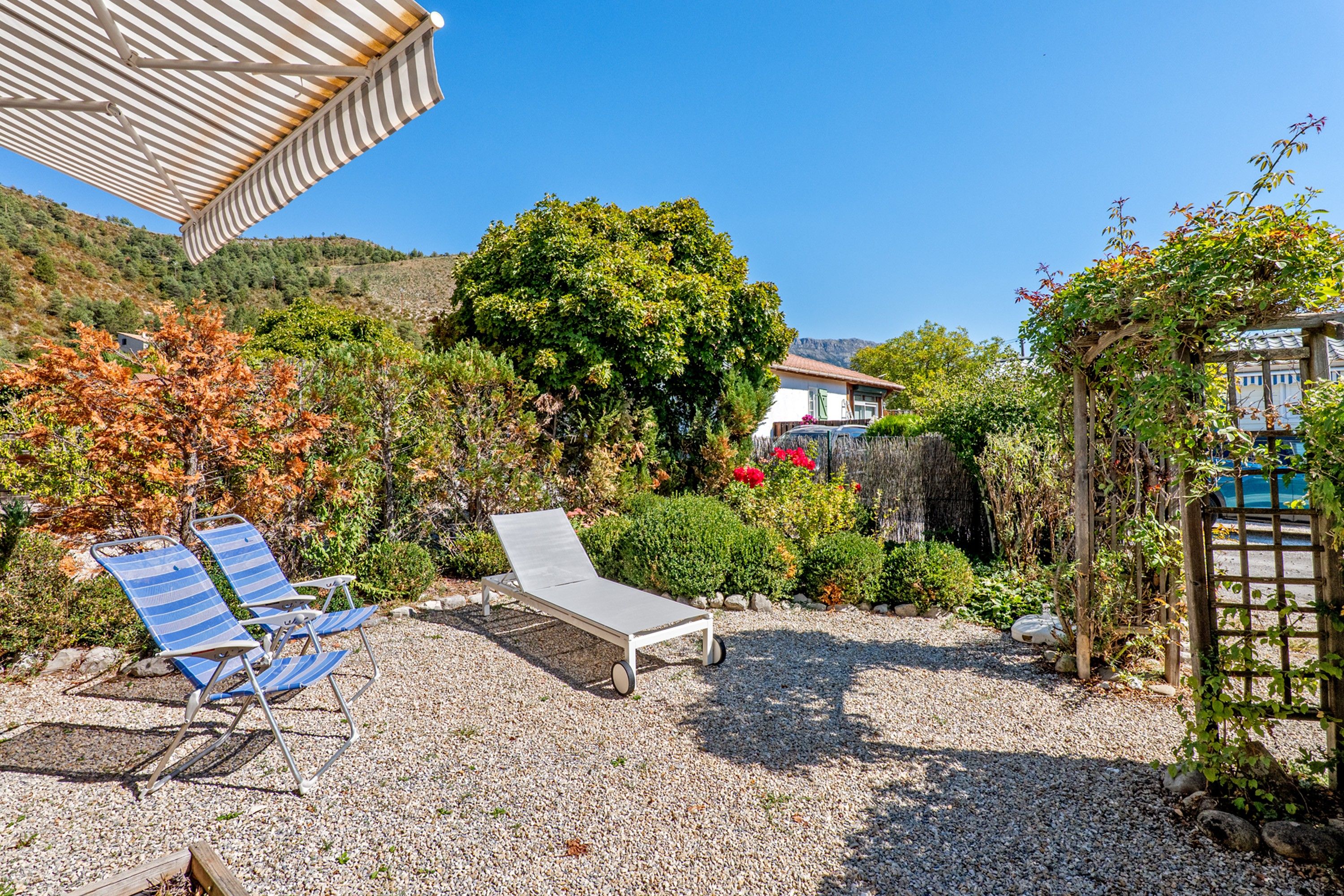 Un espace patio aménagé avec deux chaises longues et une table, entouré de gravier et de verdure.
