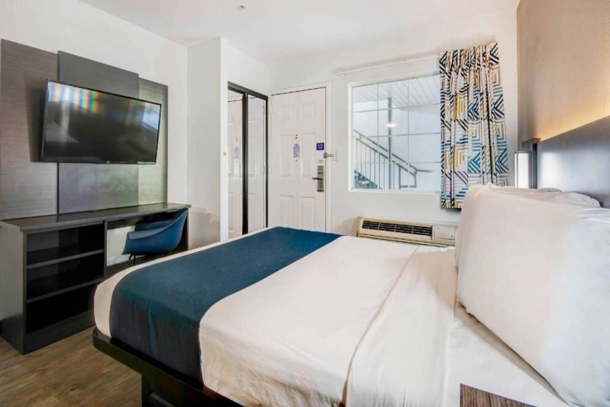 A hotel room with a bed, a desk with a chair, a television, a window with curtains, and a door.