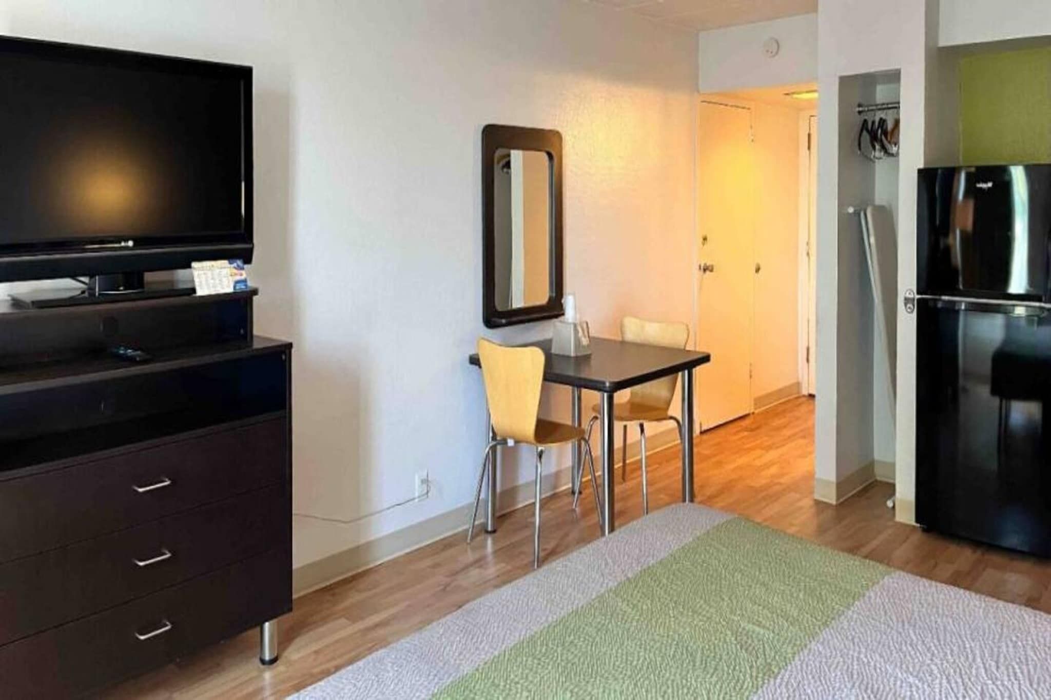 Une chambre d’hôtel équipée d’un téléviseur à écran plat, d’une petite table avec une chaise, d’un miroir, d’un réfrigérateur et d’un plancher en bois.
