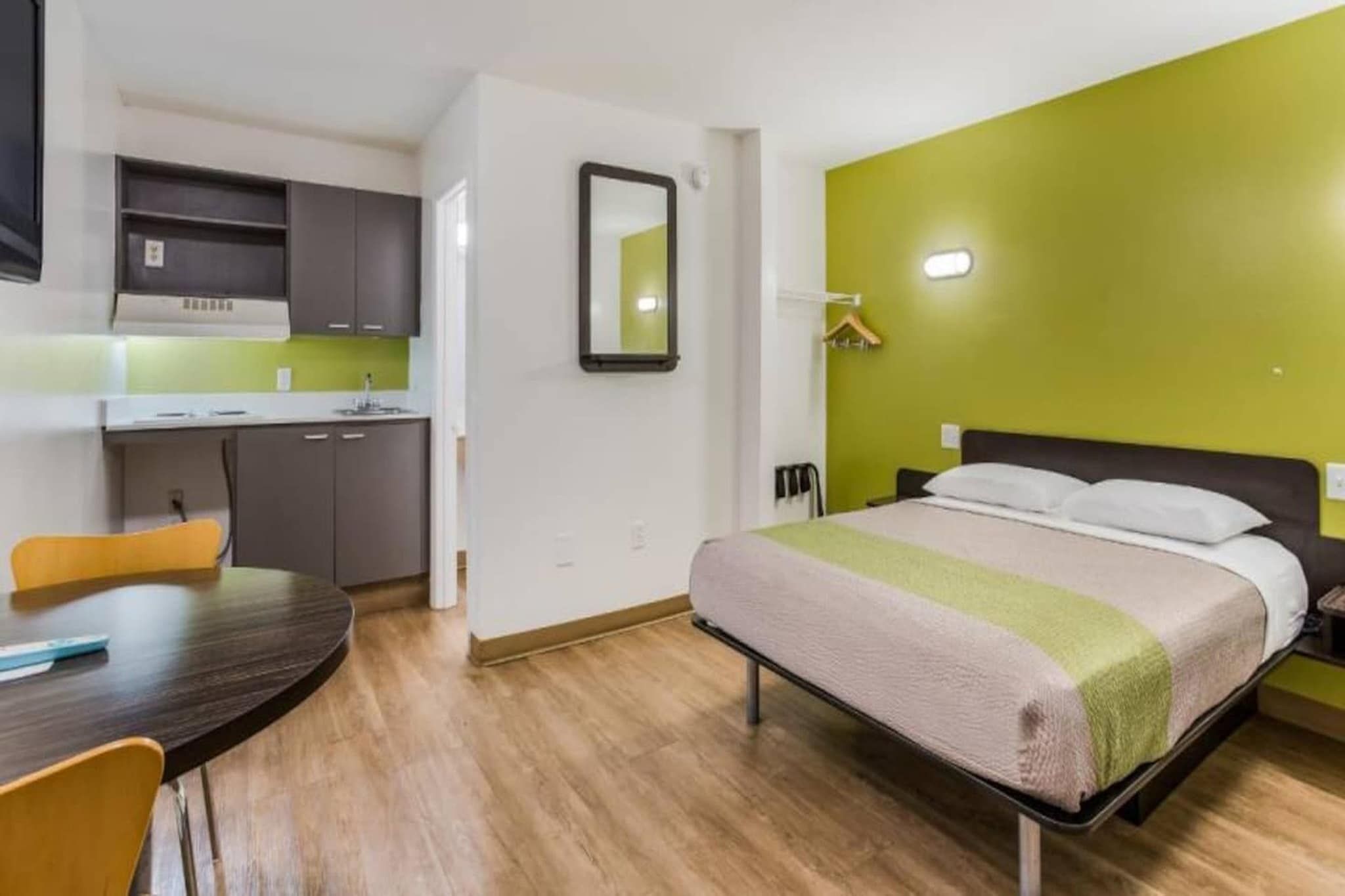 A hotel room with a bed, a desk, a chair, a mirror, and a kitchenette.