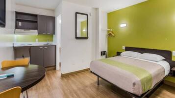 A hotel room with a bed, a desk, a chair, a mirror, and a kitchenette.
