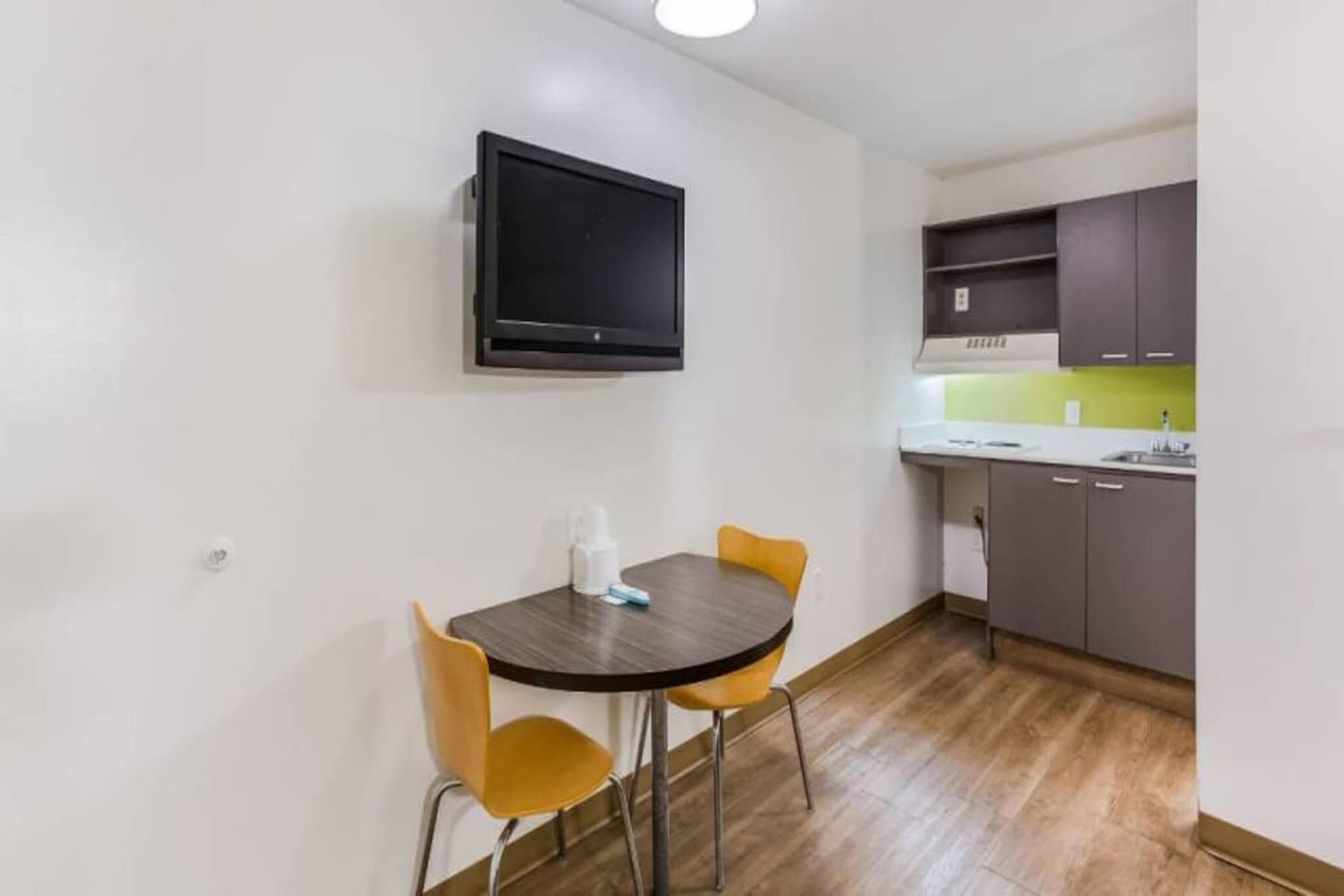 A compact kitchen with a dining area, a flat-screen TV mounted on the wall, and a small table with chairs.