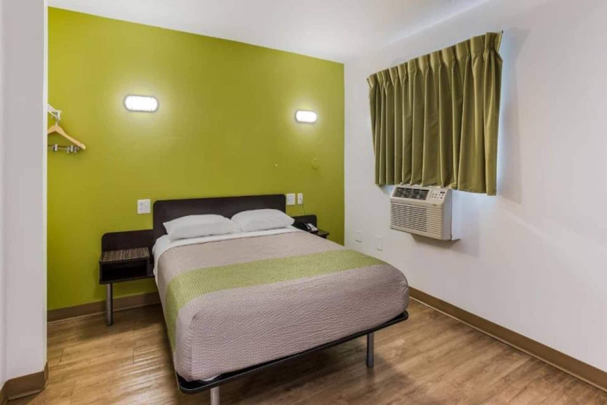 A hotel room with a bed, a nightstand, a wall-mounted air conditioner, and a window with curtains.