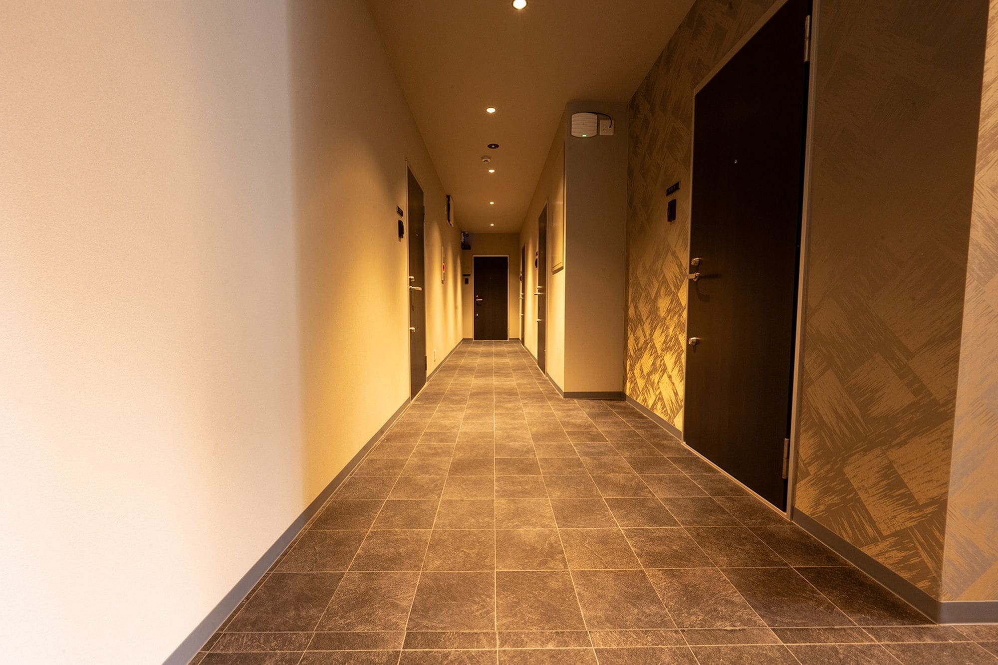 Un pasillo de hotel con suelo de baldosas, puertas a ambos lados e iluminación empotrada.