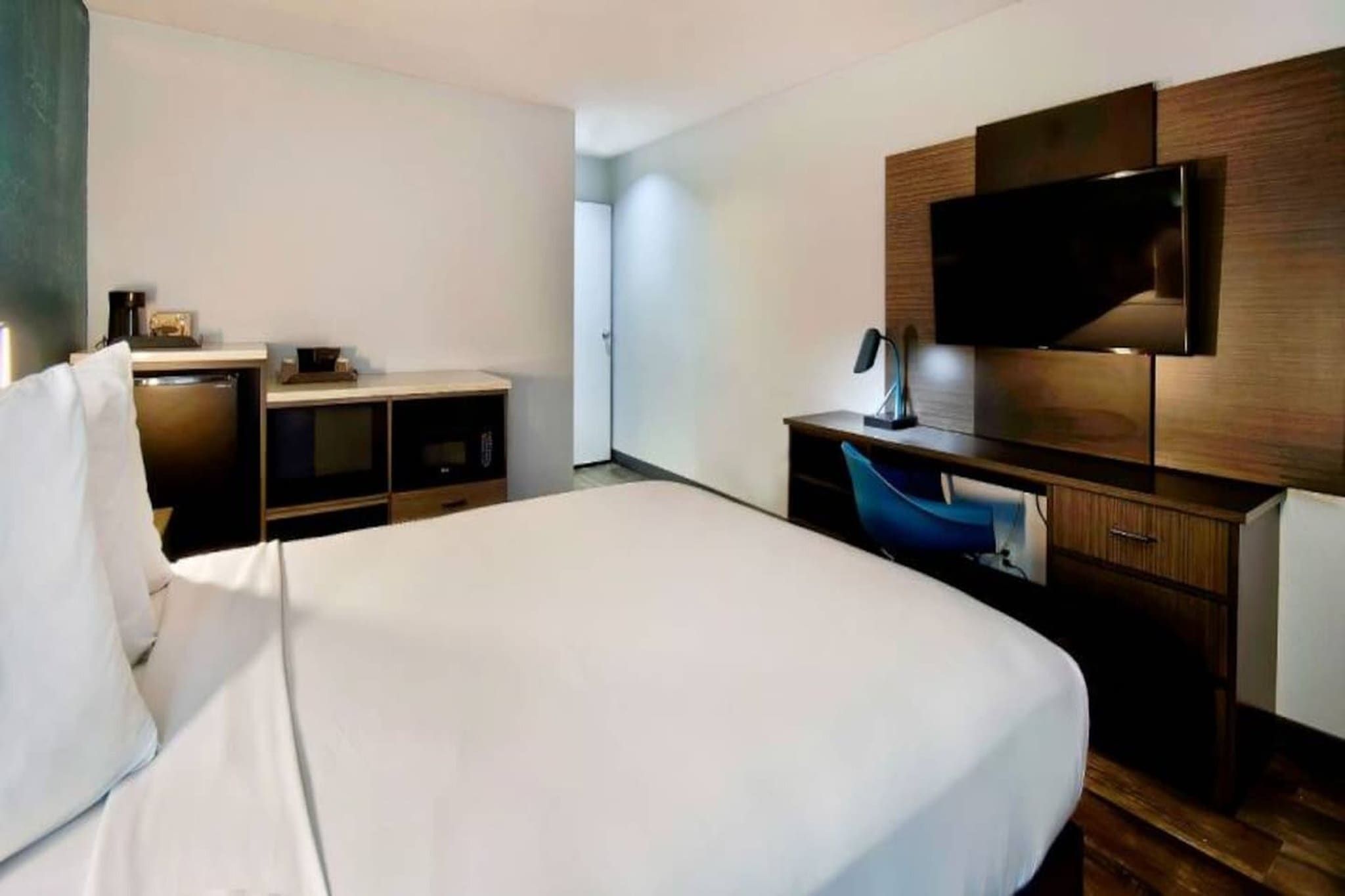 A hotel room with a large bed, a desk with a chair, a flat-screen TV mounted on the wall, and a minibar.