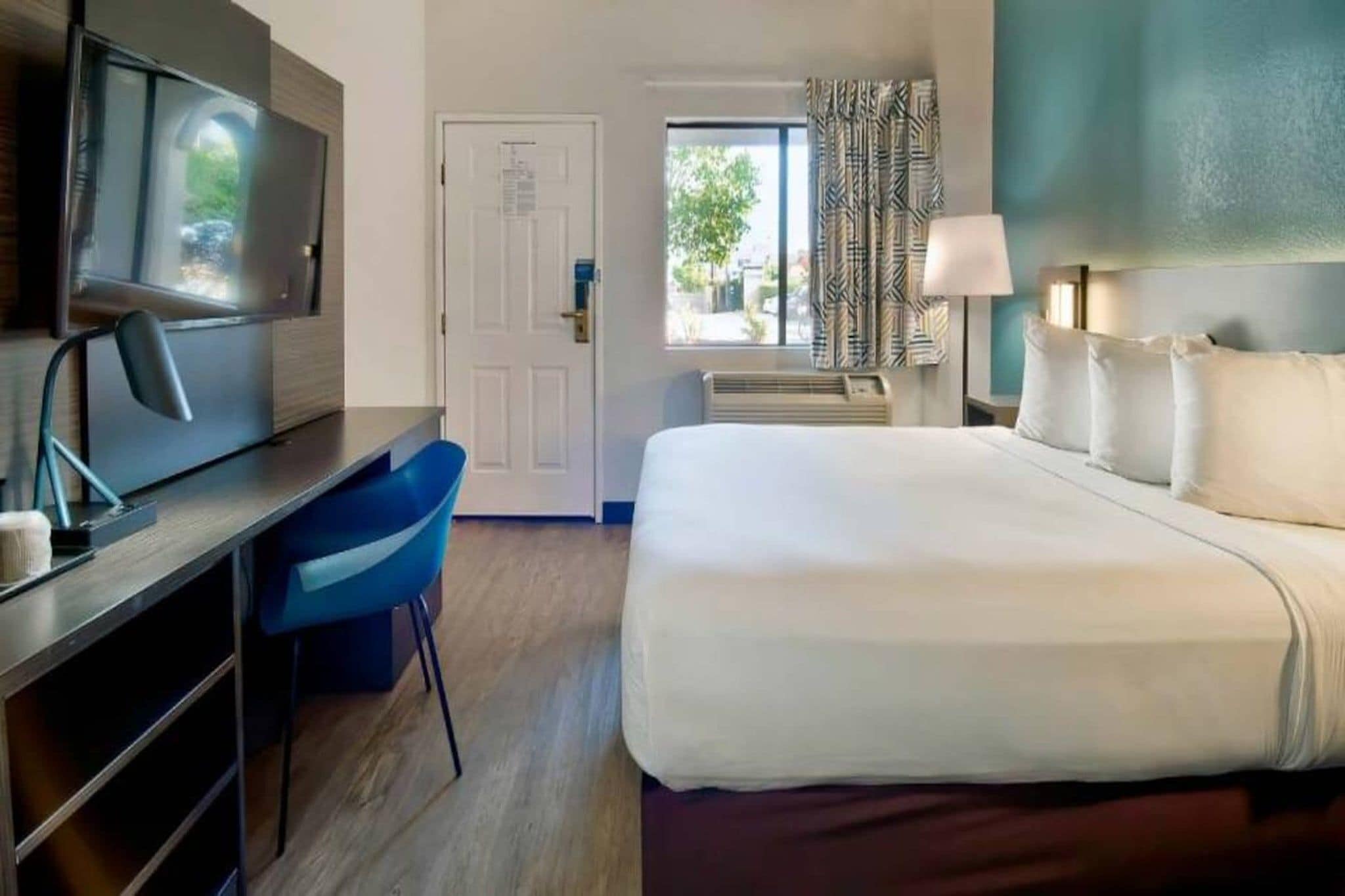 A hotel room with a bed, a desk, a chair, a television, a window with curtains, and a door.
