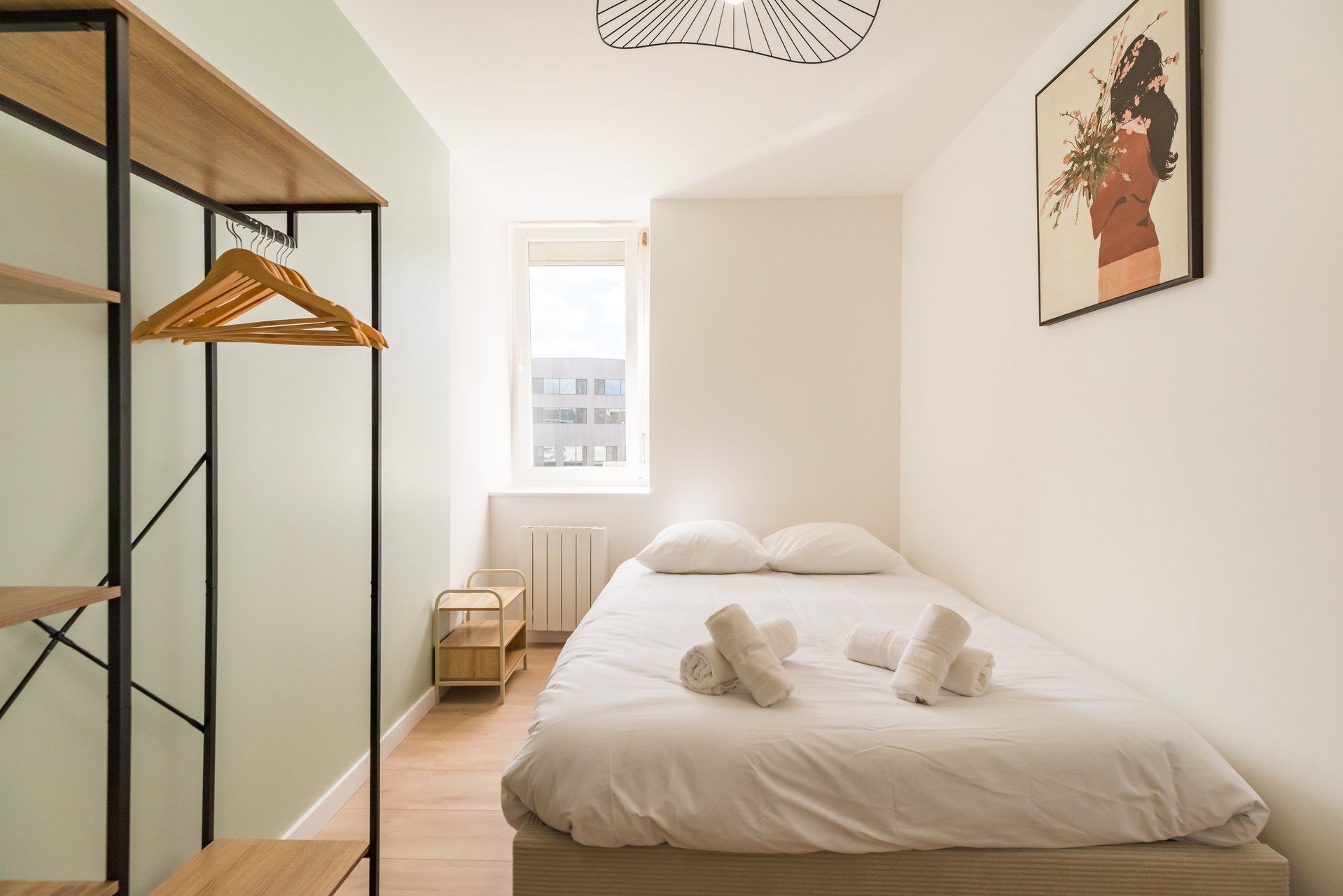Une chambre à coucher avec un lit, une armoire en bois, un radiateur et une fenêtre donnant sur des immeubles.
