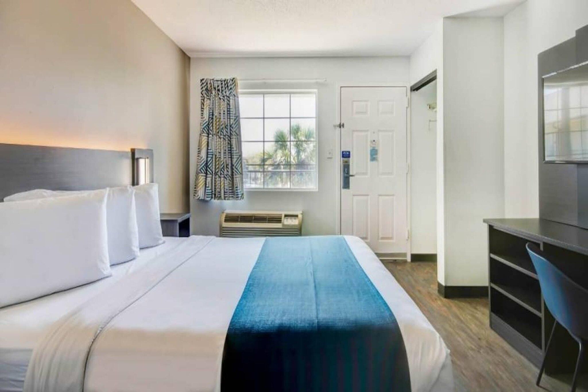 A hotel room with a bed, a desk, a chair, a window with curtains, and a door.