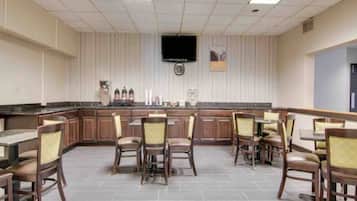 A hotel dining area with a bar, multiple tables and chairs, a flat-screen TV mounted on the wall, and a clock.