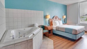 A hotel room with a large bathtub, a bed with a blue bedspread, two bedside lamps, and a view of the city through a window.