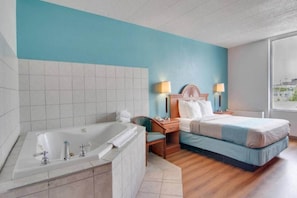 A hotel room with a large bathtub, a bed with a blue bedspread, two bedside lamps, and a view of the city through a window.