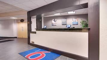 A hotel reception area with a reception desk, a blue carpet with a logo, and a wall-mounted information board.