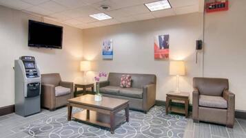Un vestíbulo de hotel con televisión de pantalla plana, cajero automático, sofá, dos sillones, una mesa de centro y alfombra estampada.